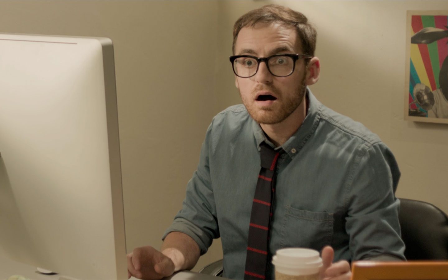 Man wearing glasses and a blue shirt with a striped tie sits at a desk, looking surprised at a large computer monitor, holding a coffee cup. | Jerad Schutt | Nashville Marketing Consultant | Fractional CMO | Jerad Schutt | Nashville Marketing Consultant | Fractional CMO