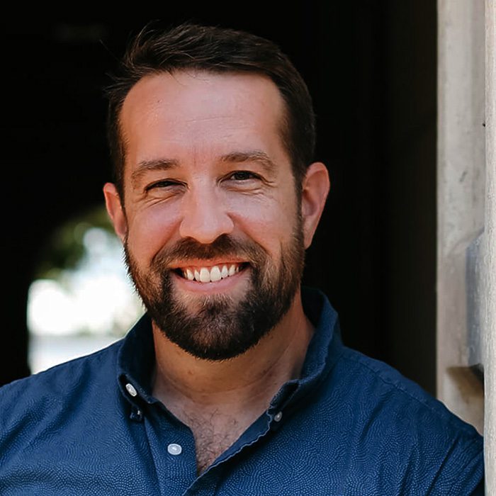 Smiling man with dark hair and a beard wearing a blue button-down shirt, standing in a doorway. | Nashville Marketing Consultant | Fractional CMO | Nashville Marketing Consultant | Fractional CMO