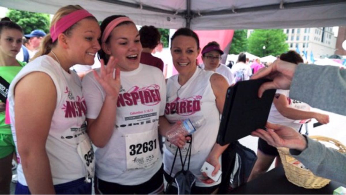 Three women in white 'Inspired' shirts pose for a photo under a canopy at an outdoor race/event, smiling and waving one hand. | Jerad Schutt | Nashville Marketing Consultant | Fractional CMO | Jerad Schutt | Nashville Marketing Consultant | Fractional CMO