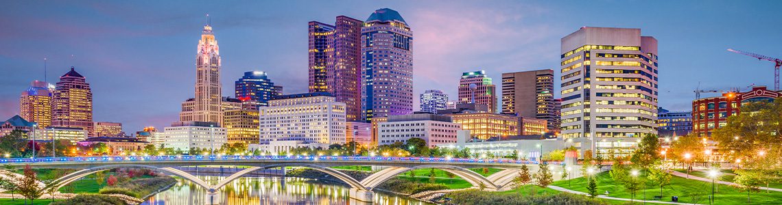 Downtown Columbus, Ohio city skyline at dusk with illuminated buildings and a lit arched bridge over a calm river ahead of manicured riverbanks. | Jerad Schutt | Nashville Marketing Consultant | Fractional CMO | Jerad Schutt | Nashville Marketing Consultant | Fractional CMO