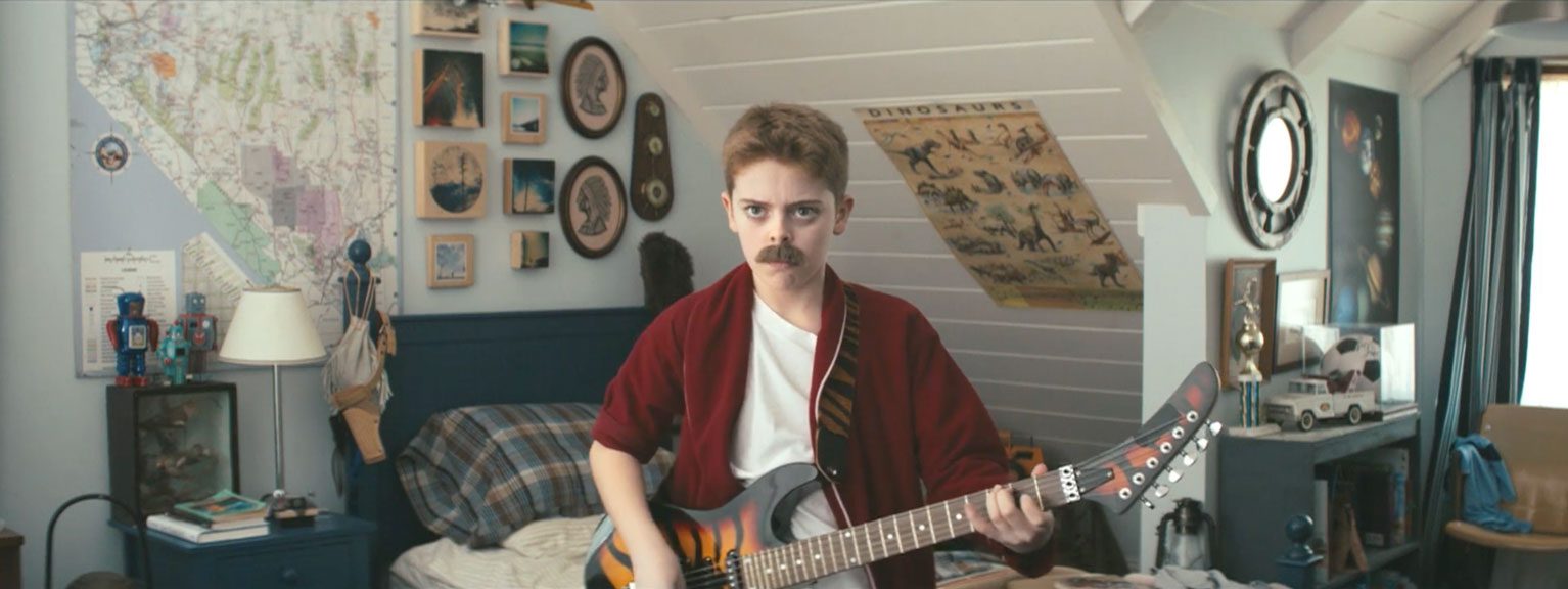 Young boy with a mustache plays an orange‑to‑black electric guitar in a cluttered bedroom filled with maps, posters, and trinkets. | Jerad Schutt | Nashville Marketing Consultant | Fractional CMO | Jerad Schutt | Nashville Marketing Consultant | Fractional CMO