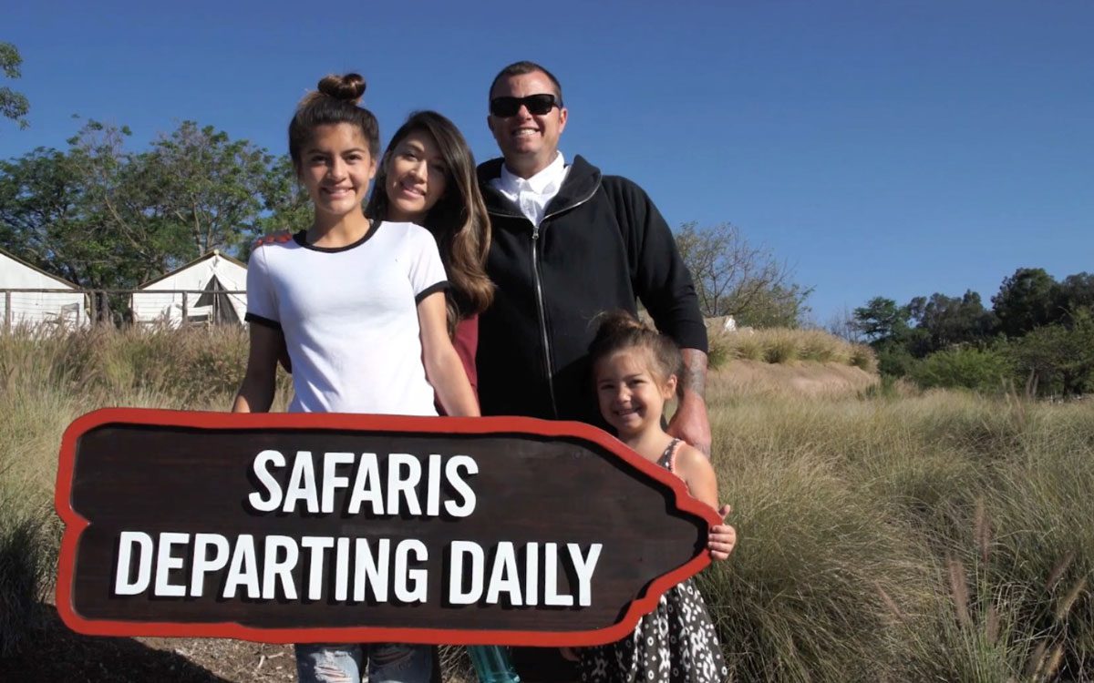 Family of four posing outdoors behind a large sign reading SAFARIS DEPARTING DAILY. | Jerad Schutt | Nashville Marketing Consultant | Fractional CMO | Jerad Schutt | Nashville Marketing Consultant | Fractional CMO