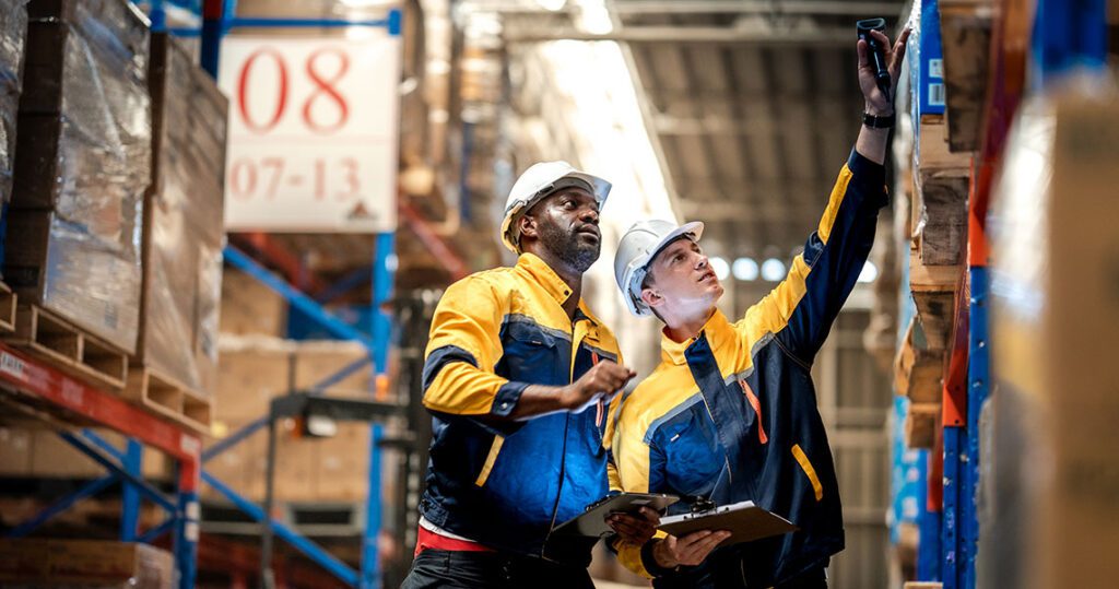Two warehouse workers in safety gear take a selfie with a smartphone while one holds a clipboard among tall shelves far in the background. | Jerad Schutt | Nashville Marketing Consultant | Fractional CMO | Jerad Schutt | Nashville Marketing Consultant | Fractional CMO