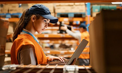 Female warehouse worker in an orange safety vest and cap typing on a laptop among shelves in a warehouse. | Jerad Schutt | Nashville Marketing Consultant | Fractional CMO | Jerad Schutt | Nashville Marketing Consultant | Fractional CMO