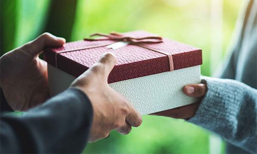 Two people exchange a red gift box with a pale base outdoors, hands near the center of the frame. | Jerad Schutt | Nashville Marketing Consultant | Fractional CMO | Jerad Schutt | Nashville Marketing Consultant | Fractional CMO