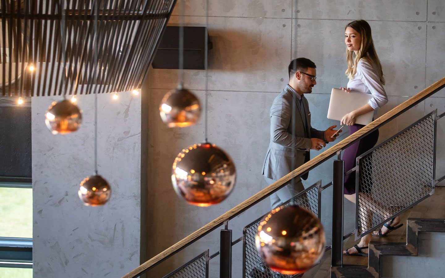 Two professionals on a modern staircase; the woman carries a laptop while the man speaks to her, mid-conversation in a business setting. | Jerad Schutt | Nashville Marketing Consultant | Fractional CMO | Jerad Schutt | Nashville Marketing Consultant | Fractional CMO