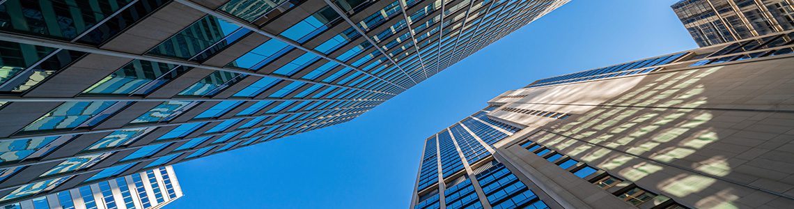 Tall glass and stone skyscrapers seen from ground level, converging toward a clear blue sky. | Jerad Schutt | Nashville Marketing Consultant | Fractional CMO | Jerad Schutt | Nashville Marketing Consultant | Fractional CMO