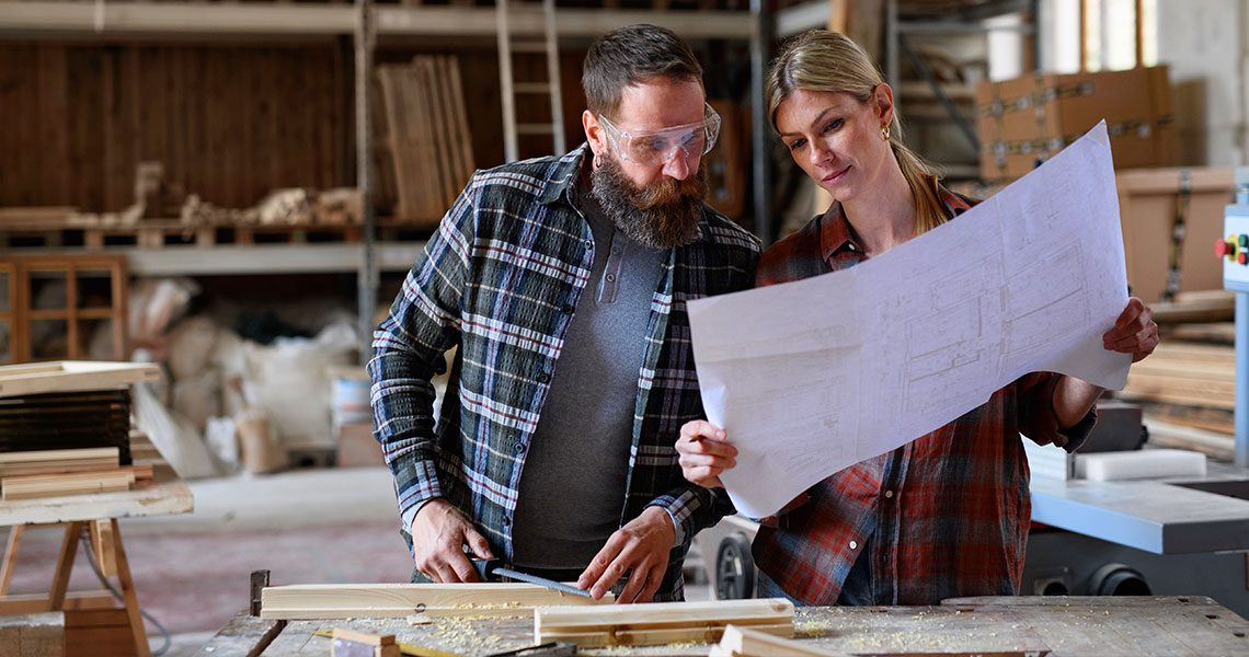 Two woodworkers in a workshop study a large blueprint together over a wooden workbench. | Jerad Schutt | Nashville Marketing Consultant | Fractional CMO | Jerad Schutt | Nashville Marketing Consultant | Fractional CMO