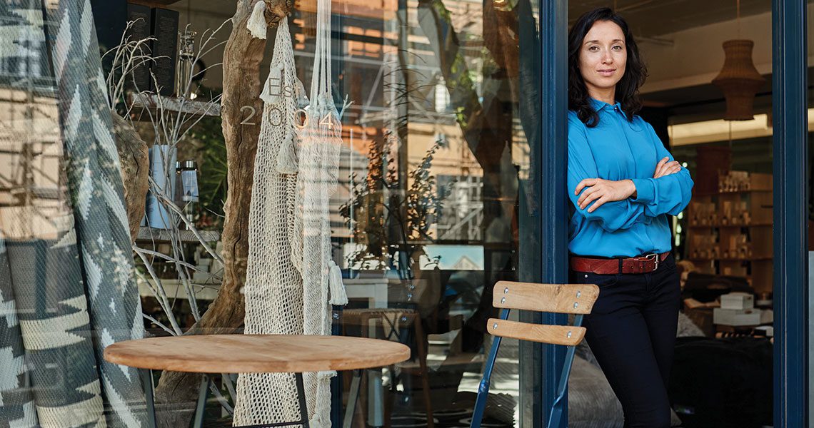 Female small business owner in a blue blouse stands with arms crossed in the doorway of a shop, with an outdoor wooden table and chair in the foreground. | Jerad Schutt | Nashville Marketing Consultant | Fractional CMO | Jerad Schutt | Nashville Marketing Consultant | Fractional CMO
