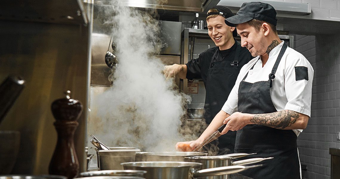 Two chefs in a busy stainless steel kitchen, one stirring pots as steam rises between them. | Jerad Schutt | Nashville Marketing Consultant | Fractional CMO | Jerad Schutt | Nashville Marketing Consultant | Fractional CMO