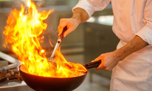 Chef in a white coat cooks with a large flame leaping from a frying pan, flipping food with a spatula. | Jerad Schutt | Nashville Marketing Consultant | Fractional CMO | Jerad Schutt | Nashville Marketing Consultant | Fractional CMO
